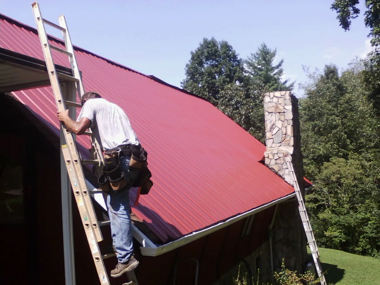 Roofer on a ladder beside a red metal roof and stone chimney — Soffit & Fascia Repair by Roofing Brothers in Casstown